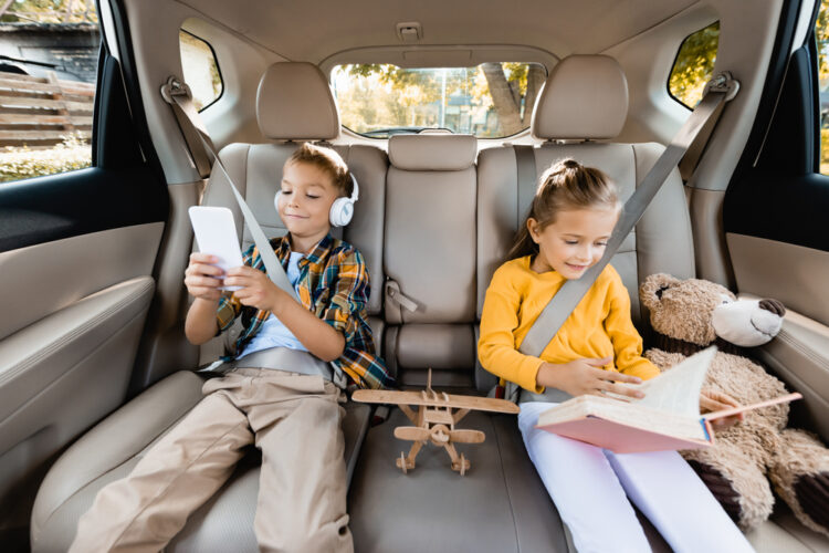 Two children sitting in back seat of car reading and playing games